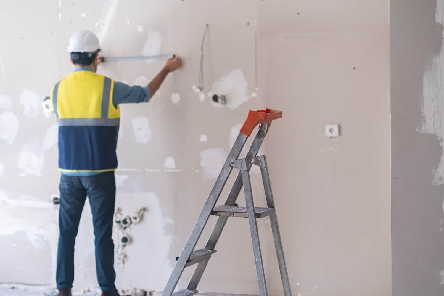 A construction worker in a hard hat and safety vest measures a wall that is under renovation,.