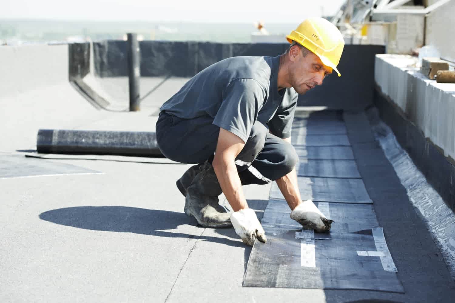 A worker performs roof waterproofing, laying a felt membrane on a flat roof.