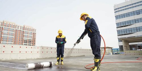 Two workers on a rooftop perform a professional roofing or waterproofing service, applying a coating with a spray gun.
